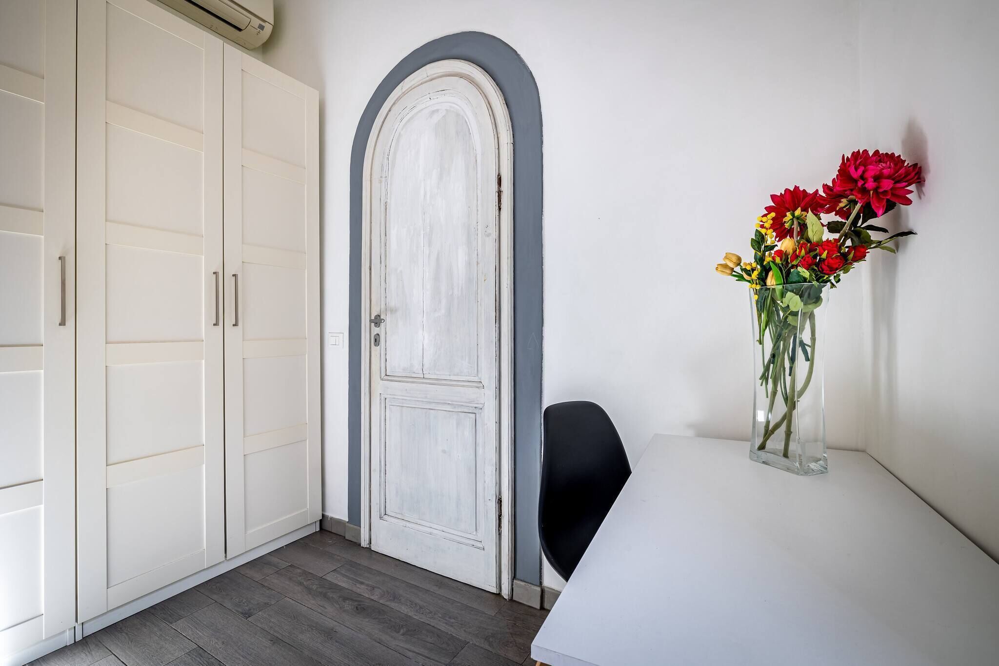Habitació amb un armari blanc, una porta blanca amb arc gris i un ram de flors sobre una taula blanca.