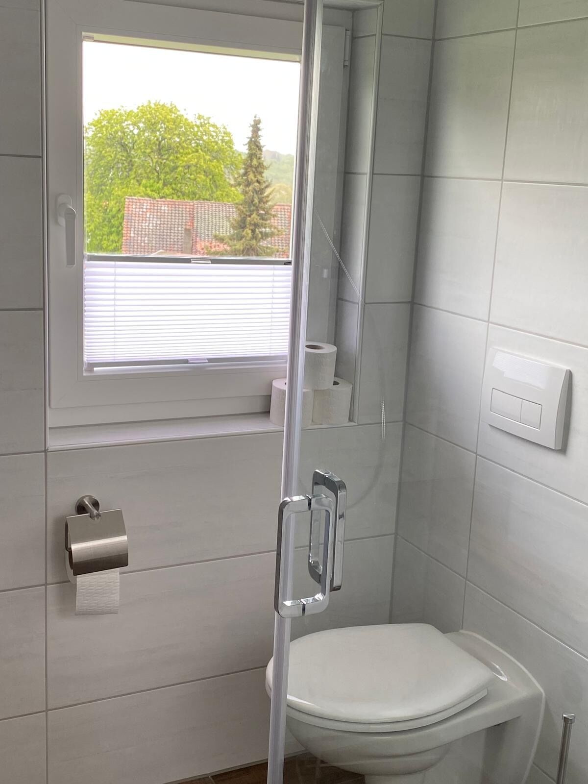 A bathroom with a toilet, a shower enclosure, a window with blinds, and a towel dispenser.