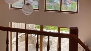 Een kamer met een schuin houten plafond, twee grote ramen met uitzicht op een kerk en huizen, en hangende bolvormige lampen.