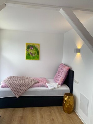 Une chambre à coucher comprenant un lit, un coussin décoratif, un plaid, un tableau accroché au mur et une lampe sur pied.