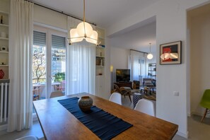 Ein Essbereich mit einem Holztisch, einer Vase und einer schwarzen Tischmatte. Im Hintergrund ist ein Fernseher zu sehen und ein Stuhl mit einem grünen Sitzkissen.