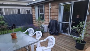A wooden deck with a table and white plastic chairs, a covered grill, and potted plants. - North east valley apartment (Dunedin)