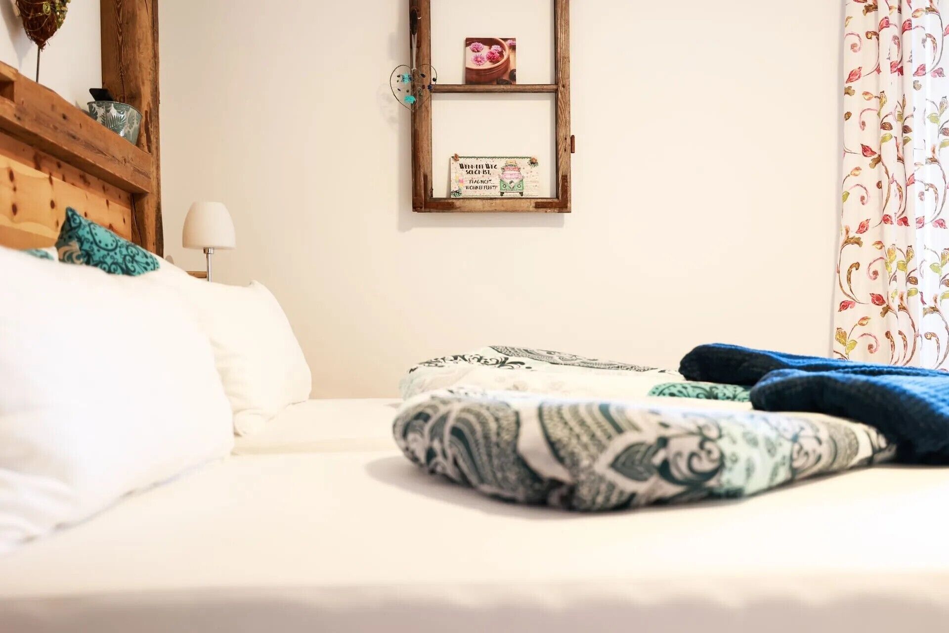 A bed with a white comforter and a patterned blanket, a wooden headboard, a bedside table with a lamp, and a wall-mounted shelf with decorative items.