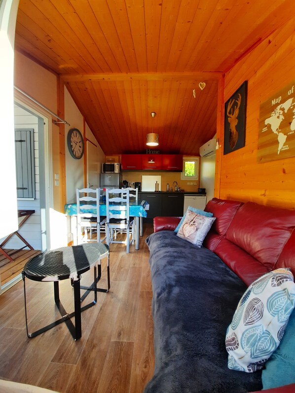 A cozy cabin interior with a wooden ceiling, a dining area with a table and chairs, a sofa with cushions, and a kitchen area with appliances.
