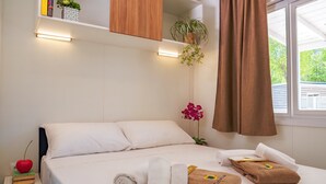 Una cama bien hecha con sábanas blancas, una mesita de noche con un libro y una manzana roja, y una ventana con cortinas marrones.