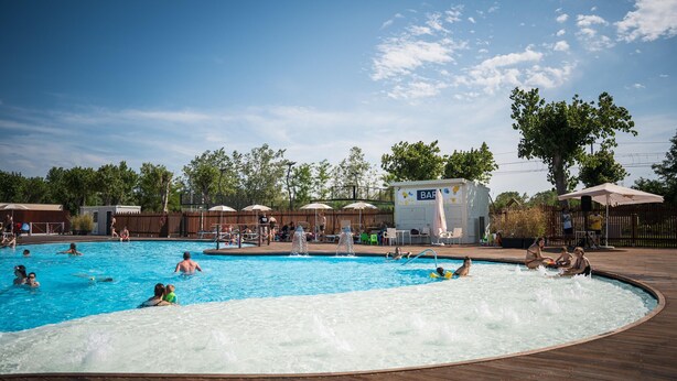 Una piscina con gente, una fuente de agua y una zona cubierta con sombrillas.