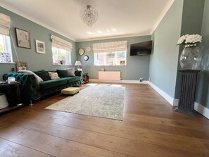 A living room with a green sofa, a wooden floor, a chandelier, a fireplace, and a TV mounted on the wall. - Spacious 4-Bedroom Coastal Retreat in Pett, East Sussex (Pett)