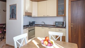 A kitchen with beige cabinets, a stove, sink, and a microwave. There is a dining table with chairs and a bowl of fruit. - Gatto errante (Pesaro)