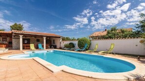 Ein Pool mit geschwungener Kante, umgeben von einer gefliesten Terrasse. Im Hintergrund sind mehrere Liegestühle und ein Gebäude mit steinernen Wänden und einem gefliesten Dach zu sehen.