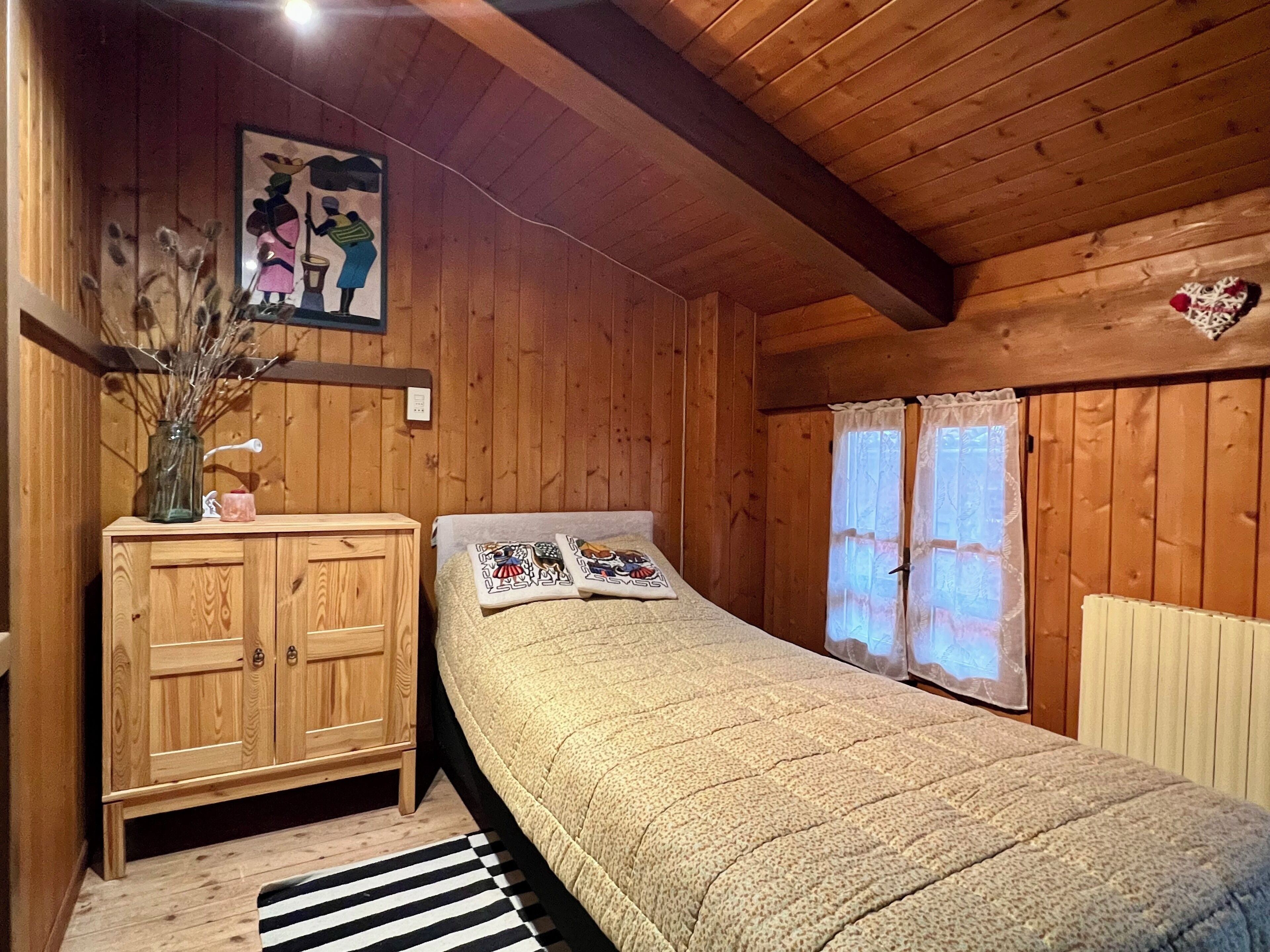 A wooden-paneled room with a bed, a wooden cabinet, a vase with dried flowers, and a painting on the wall.