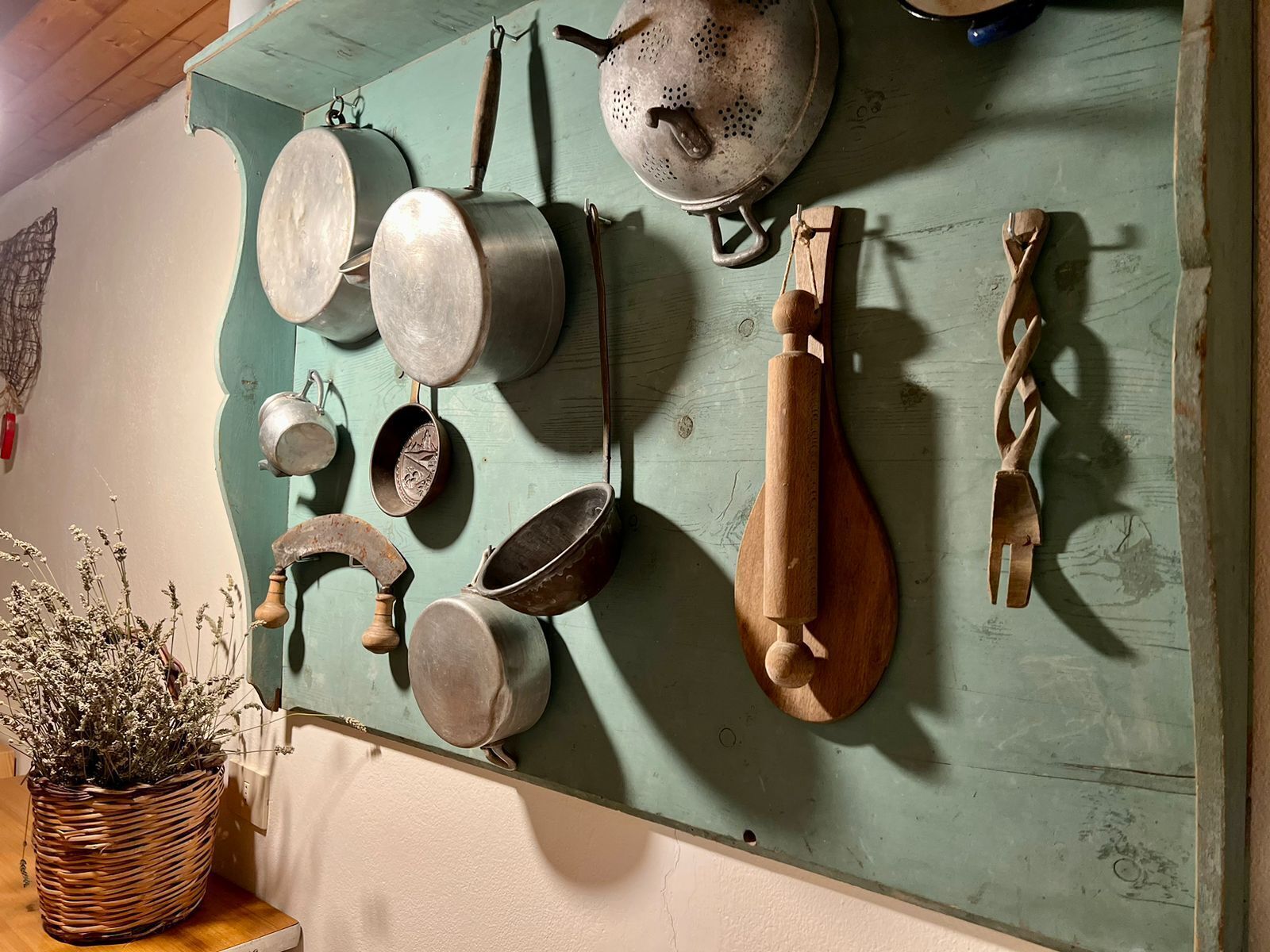 Una cucina con diverse pentole e padelle appese al muro, un mattarello di legno e un cesto di vimini con fiori secchi.