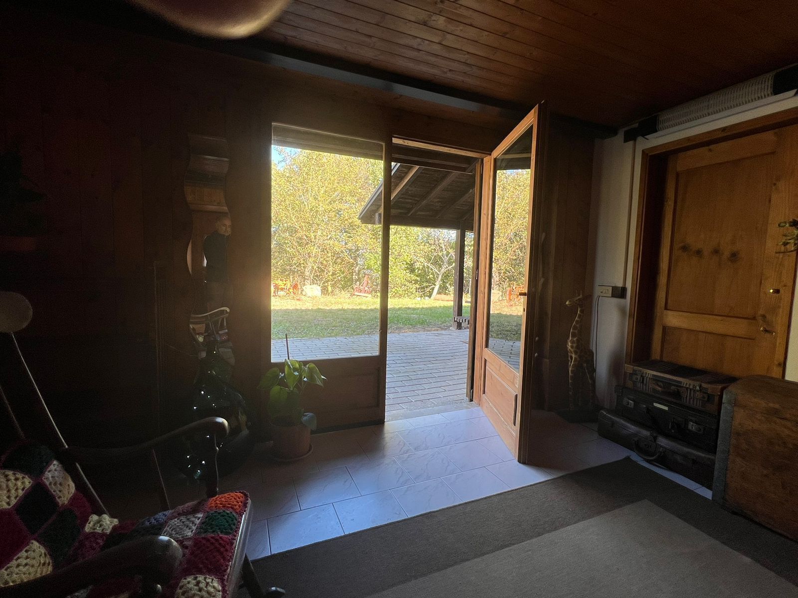 A cozy room with a wooden ceiling, a large open door leading outside, a wooden bench, and a small table with a plant.