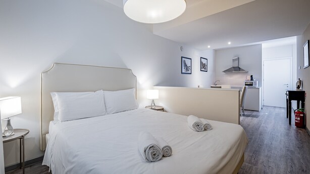 Ett hotellrum med en stor säng, ett nattduksbord med en lampa, ett litet kök och en dörr som leder till ett annat rum.