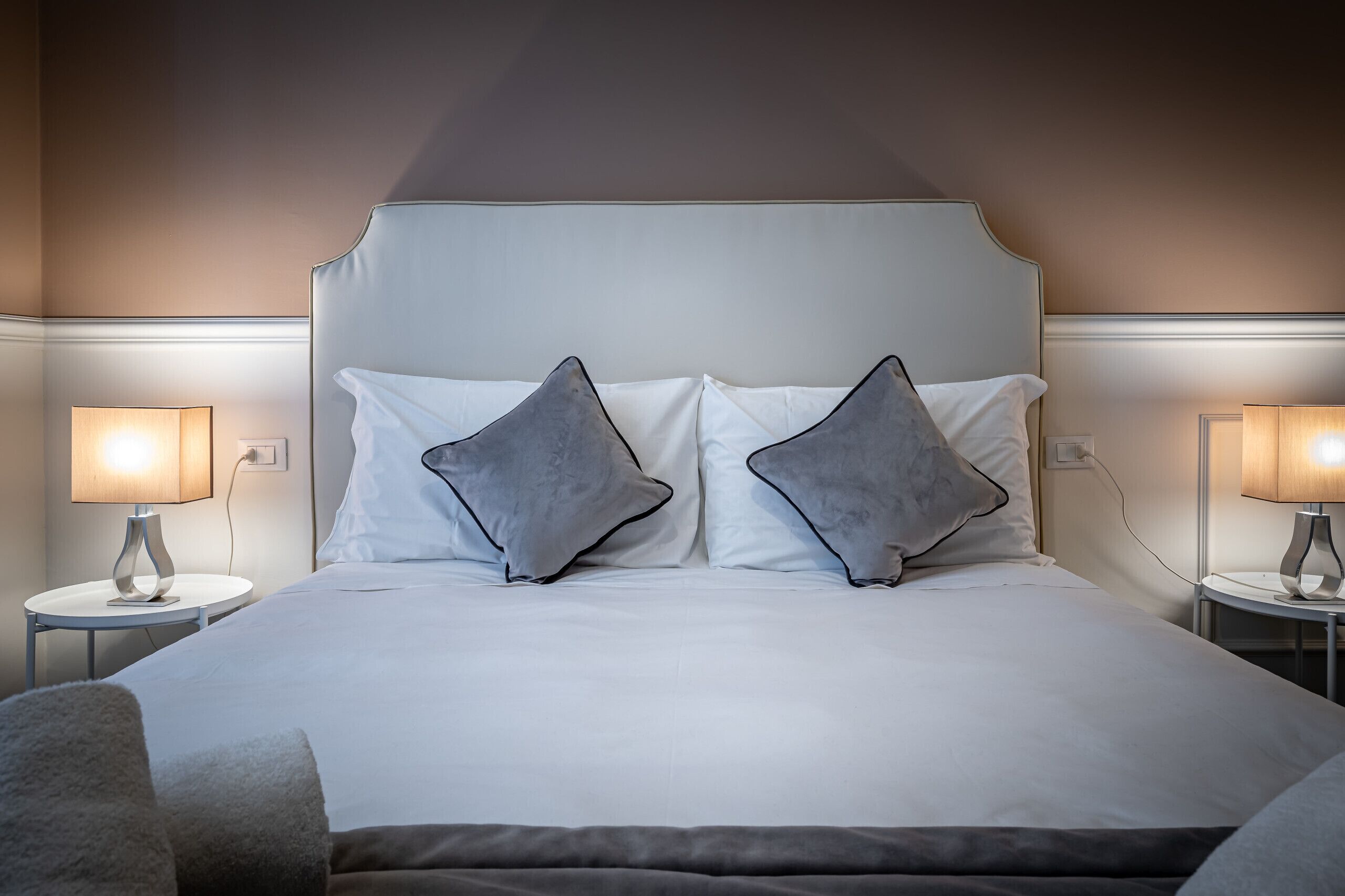 Un lit bien fait, avec du linge de lit blanc et deux oreillers gris, flanqué de deux tables de chevet sur lesquelles sont posées des lampes.