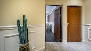 Un pasillo con una puerta de madera, un jarrón de vidrio con un cactus, y una vista parcial de una habitación con una mesa y sillas blancas.