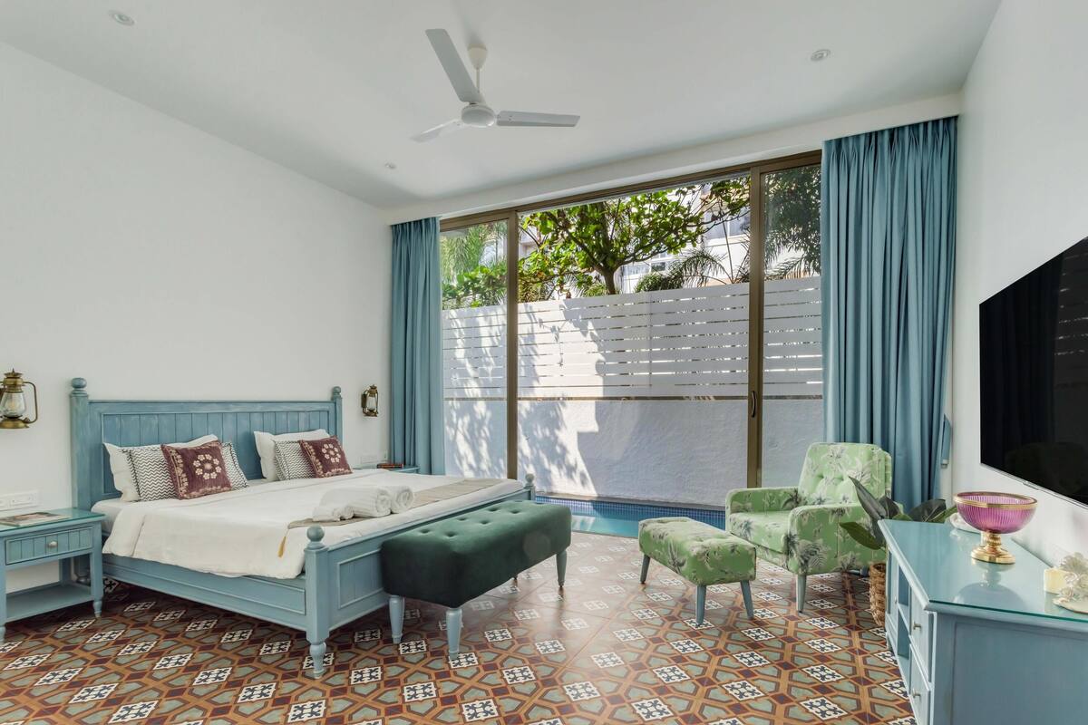 A bedroom with a bed, a bench, a chair, a TV, and a window with curtains.