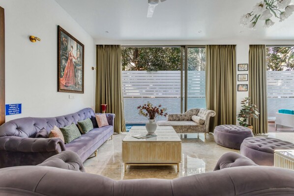 A living room with a purple sofa, a wooden coffee table, and a white armchair. There is a painting on the wall and a sign on the left. - Nerul Nirvana Nest by Dia Hotels (Nerul)