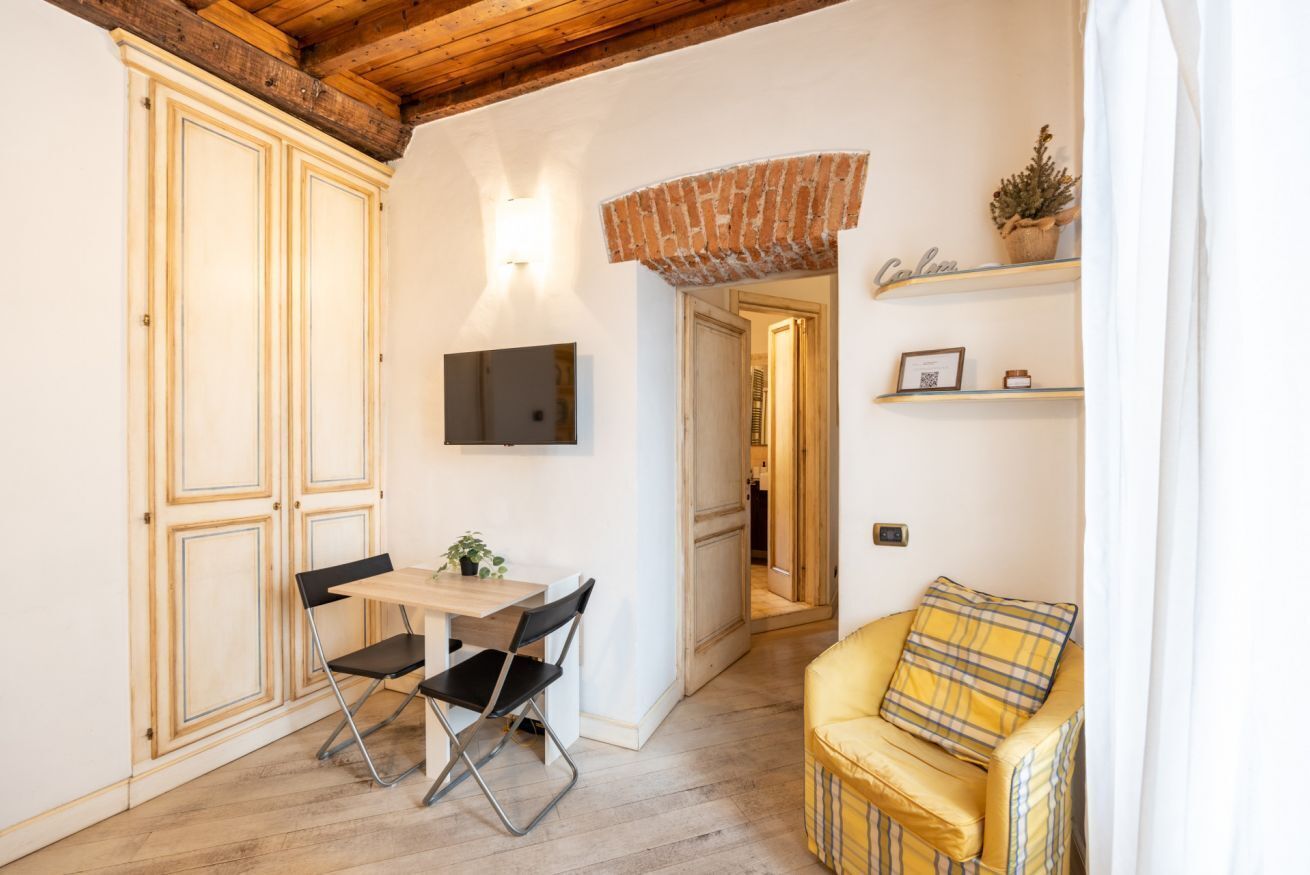 Une pièce de taille modeste comprenant une table à manger, des chaises, un fauteuil jaune, un téléviseur fixé au mur et une arche en briques donnant sur une autre pièce.