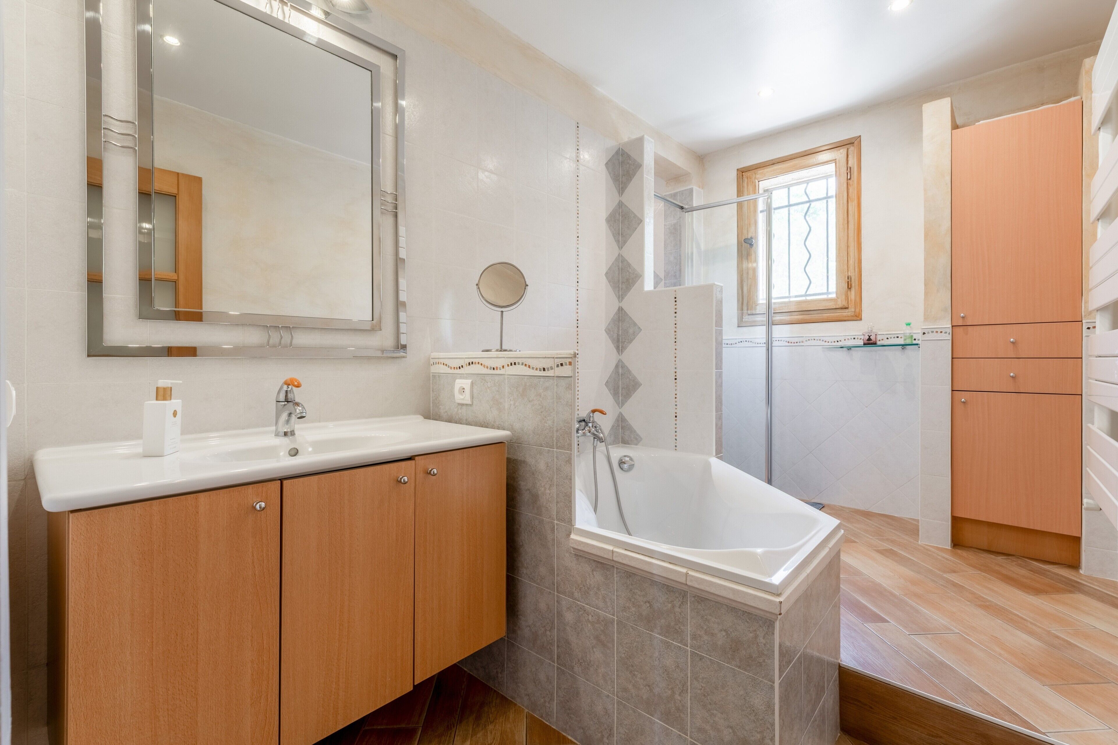 Ein modernes Badezimmer mit einer Badewanne, einem großen Spiegel, Holzschränken und einem Fenster.