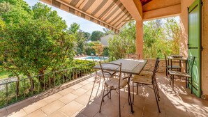 Une terrasse couverte avec une table et des chaises, une piscine en arrière-plan et une végétation luxuriante.