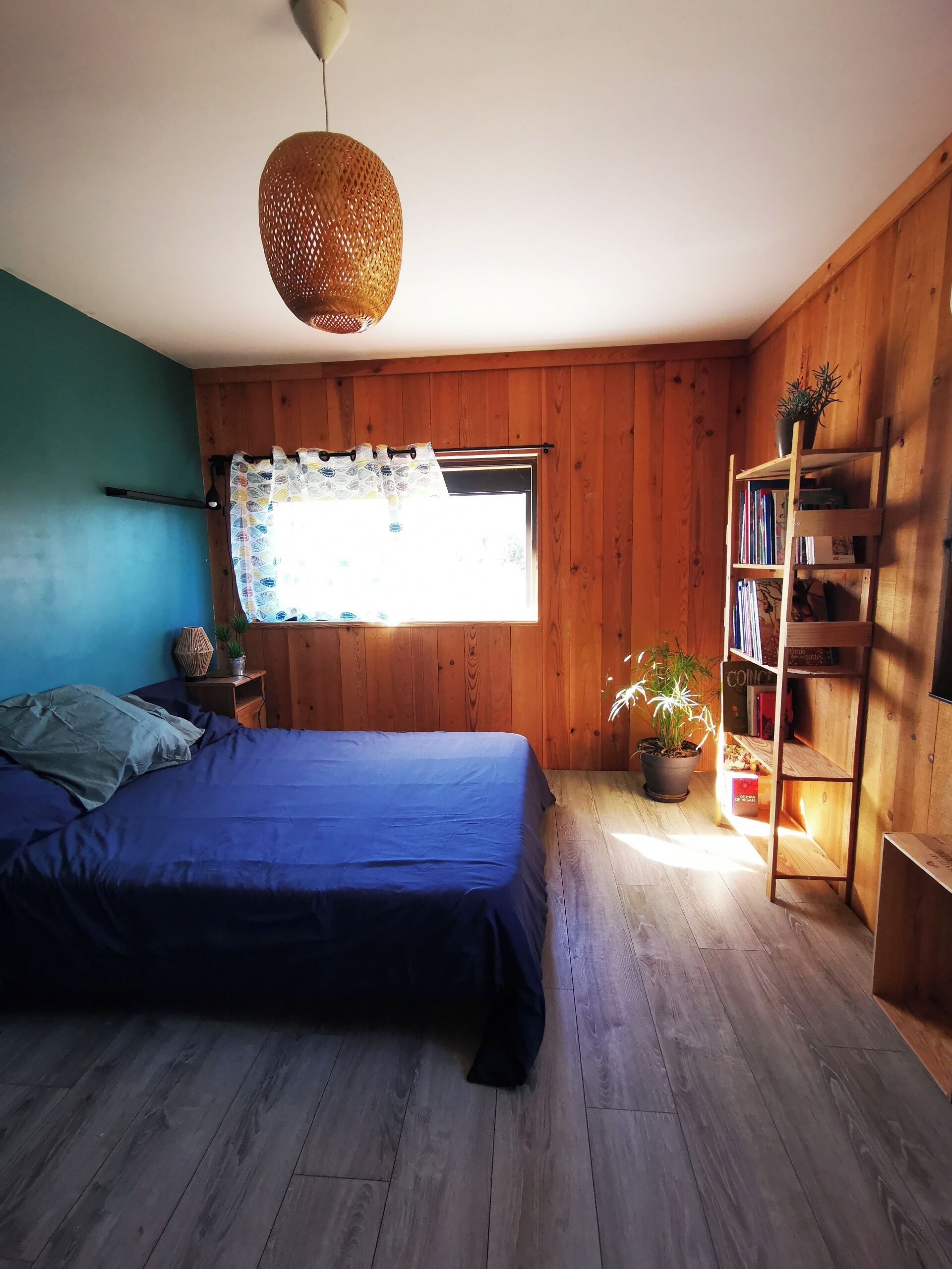 Un dormitorio con cama, ventana con cortinas, estantería de madera y lámpara colgante.