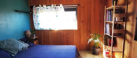 Un dormitorio con cama, ventana con cortinas, estantería de madera y lámpara colgante.
