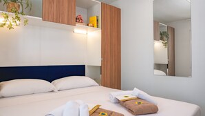 A hotel room with a bed, towels, a mirror, and a shelf with plants and books. - Friendly Mobile Home in Rimini Family Village (Rimini)