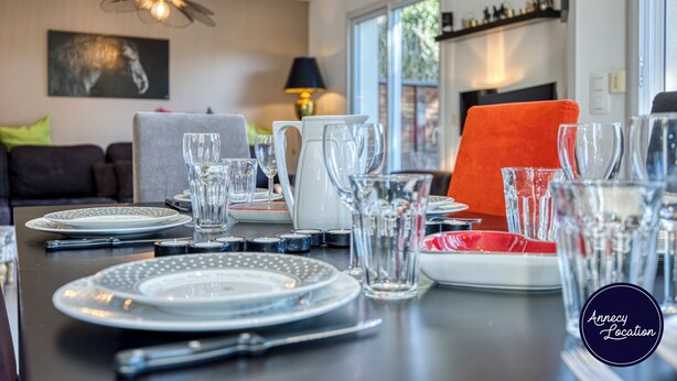 Een eettafel gedekt met borden, glazen en bestek, met een bank en lamp op de achtergrond.