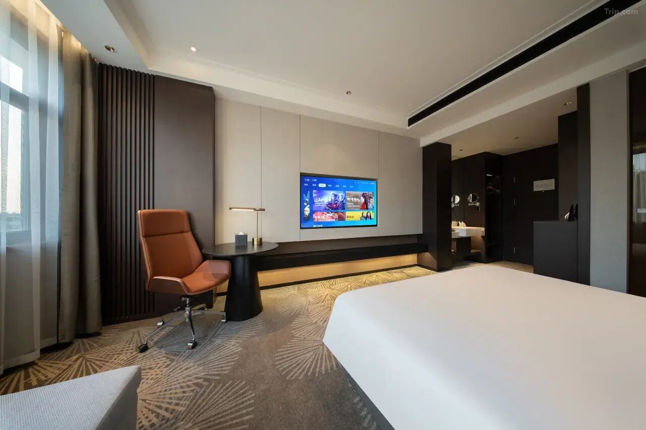 Ein modernes Hotelzimmer mit einem großen Bett, einem Schreibtisch mit Stuhl, einem flachbildfernseher an der Wand und einem großen Fenster mit Vorhängen.