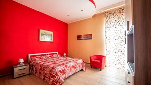 Une chambre avec une couvre-lit à motifs rouges et blancs, une chaise rouge, une table de chevet avec une lampe et un tableau au mur.