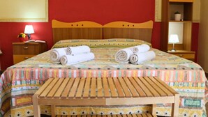Ein Hotelzimmer mit einem Bett, zwei gerollten Handtüchern, einer hölzernen Bank, einem Nachttisch mit Lampe und einem Regal mit einer Pflanze.