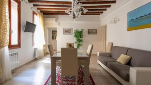 Uma sala de estar com sofá, mesa de jantar com cadeiras, lustre e um quadro na parede.