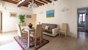 Uma sala de estar com área de jantar, vigas de madeira no teto, sofá, duas poltronas, mesa com cadeiras, tapete com padrão preto e vermelho e um quadro na parede.