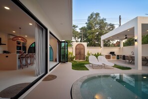 Un área de piscina moderna al aire libre con sillones de descanso, una zona de barbacoa y una cocina con paredes de vidrio.