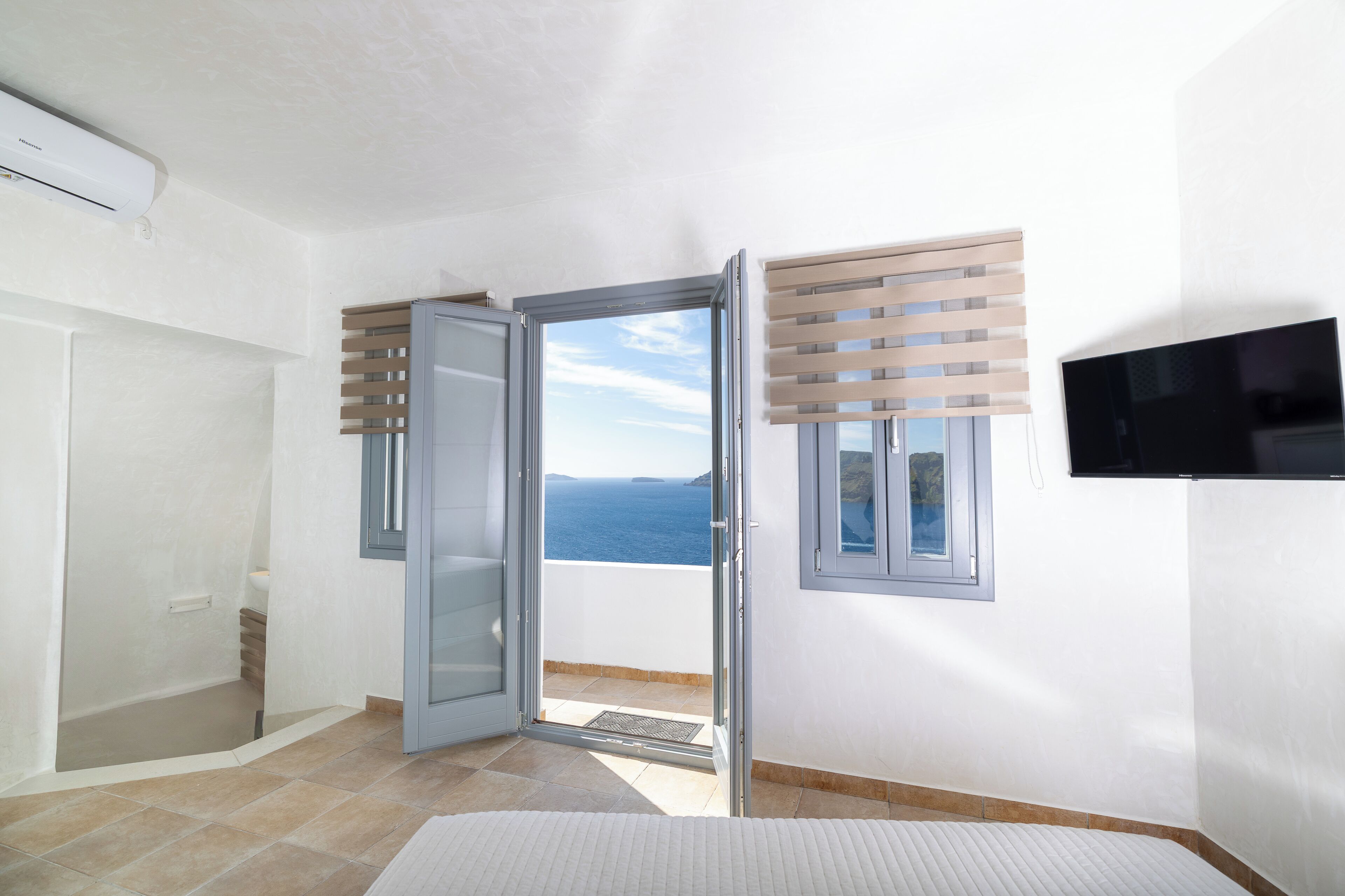 A room with a sliding glass door leading to a balcony with a sea view, a built-in air conditioning unit, and a television mounted on the wall.