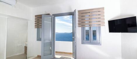 A room with a sliding glass door leading to a balcony with a sea view, a built-in air conditioning unit, and a television mounted on the wall.