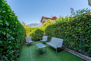 A patio with two metal chairs and a small table, surrounded by lush green hedges and a mountain view in the background. - Bellagio Breva - One Bedroom Apartment, Sleeps 2 (Bellagio)