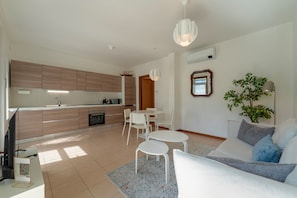 Eine moderne Küche mit Holzschränken, ein Essbereich mit weißen Stühlen, ein Sofa mit blauen Kissen und eine Topfpflanze.