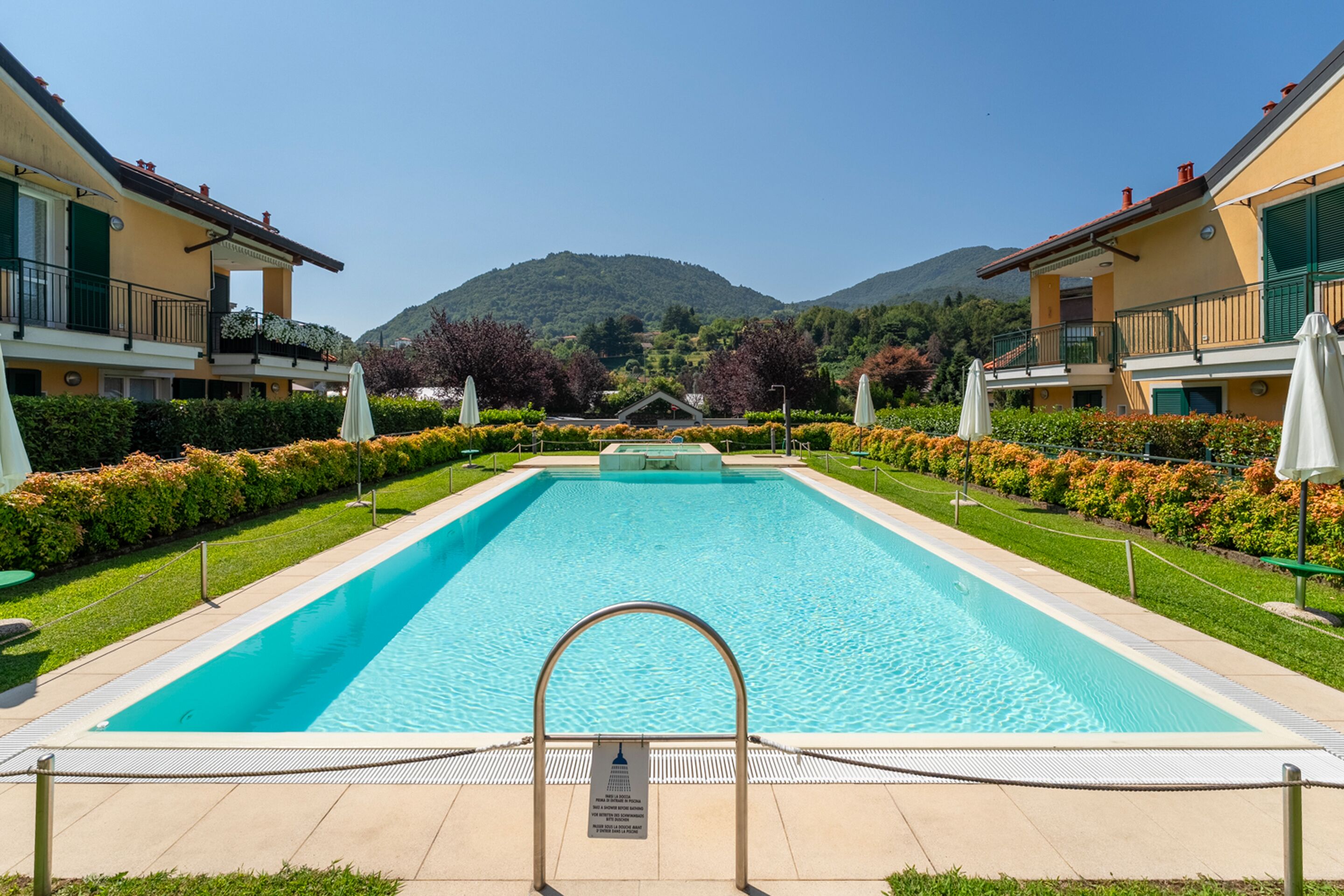 Piscina rectangular con una escalera metálica en primer plano, rodeada de setos bien cuidados y una hilera de edificios con balcones.