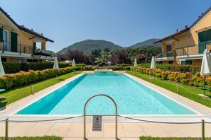 A rectangular swimming pool with a metal ladder in the foreground, surrounded by neatly trimmed hedges and a row of buildings with balconies. - Bellagio Breva - One Bedroom Apartment, Sleeps 2 (Bellagio)