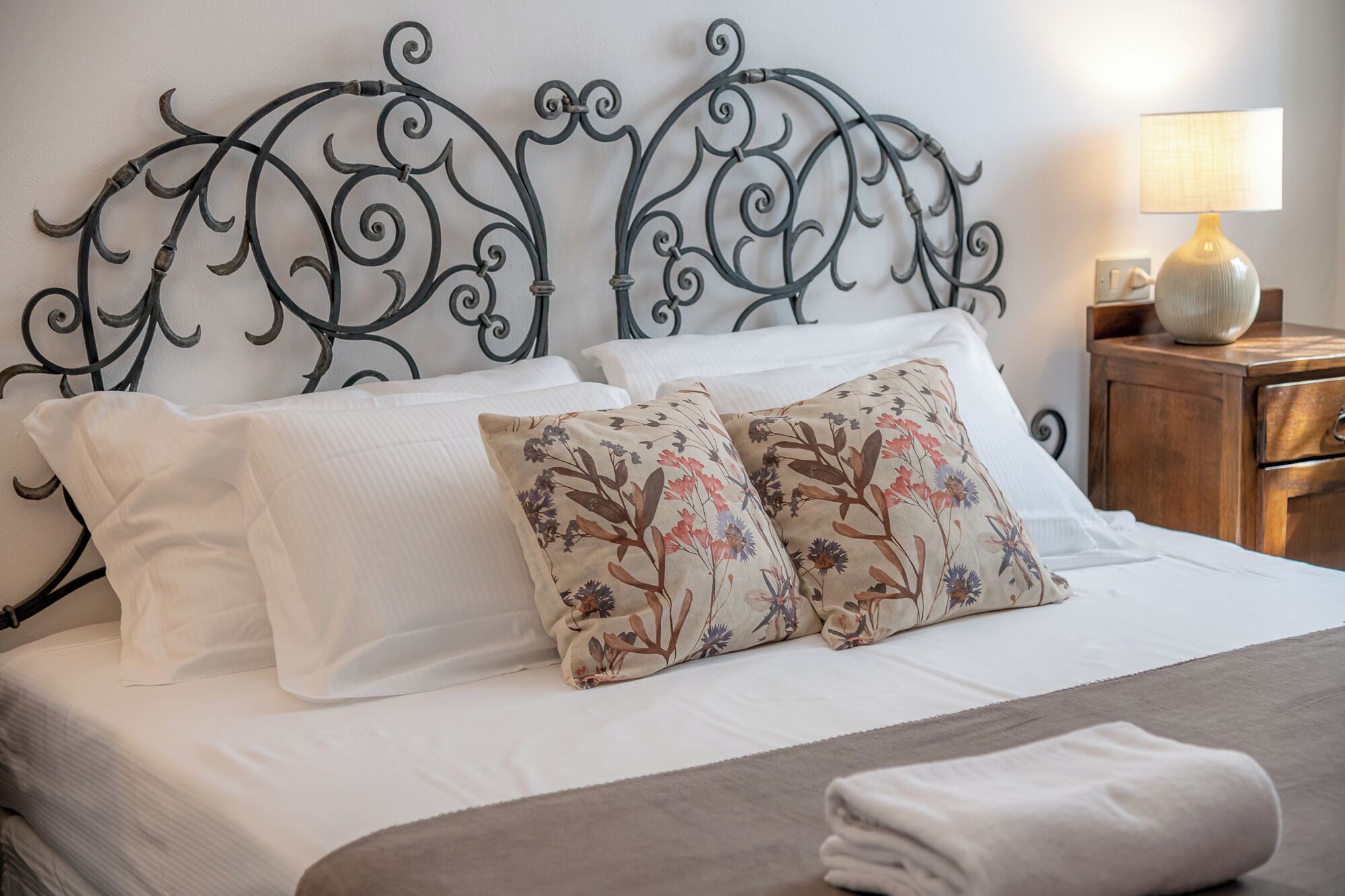 Una cama bien hecha con sábanas blancas, dos almohadas decorativas con estampado floral y un cabecero de metal con diseños intrincados.