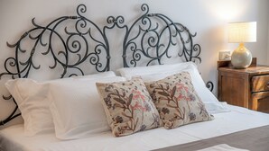Ein ordentlich bezogenes Bett mit weißen Leinentextilien, zwei dekorative Kissen mit floralem Muster und ein Metallkopfteil mit aufwendigen Verzierungen.