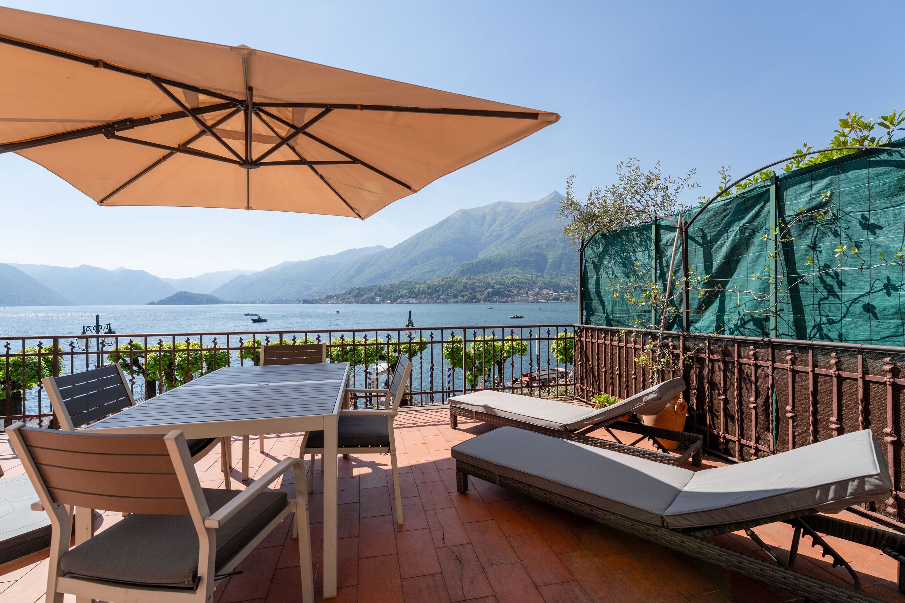 A terrace with a table, chairs, a lounge chair, and an umbrella overlooking a lake with mountains in the background.
