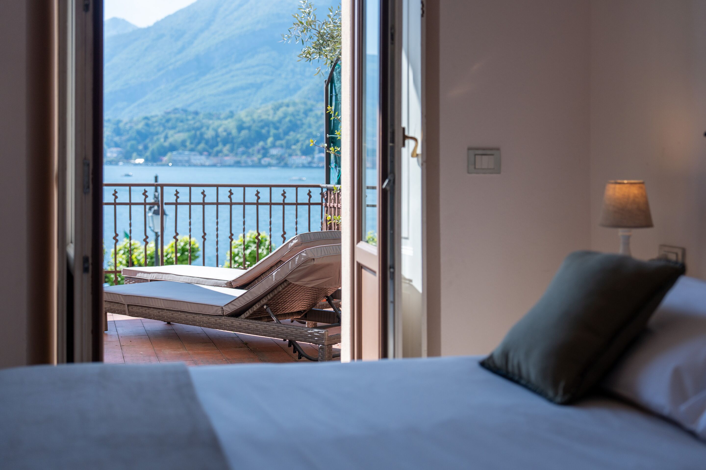 A bedroom with a bed, a nightstand, a lamp, and a view of a balcony with a railing and a chair.
