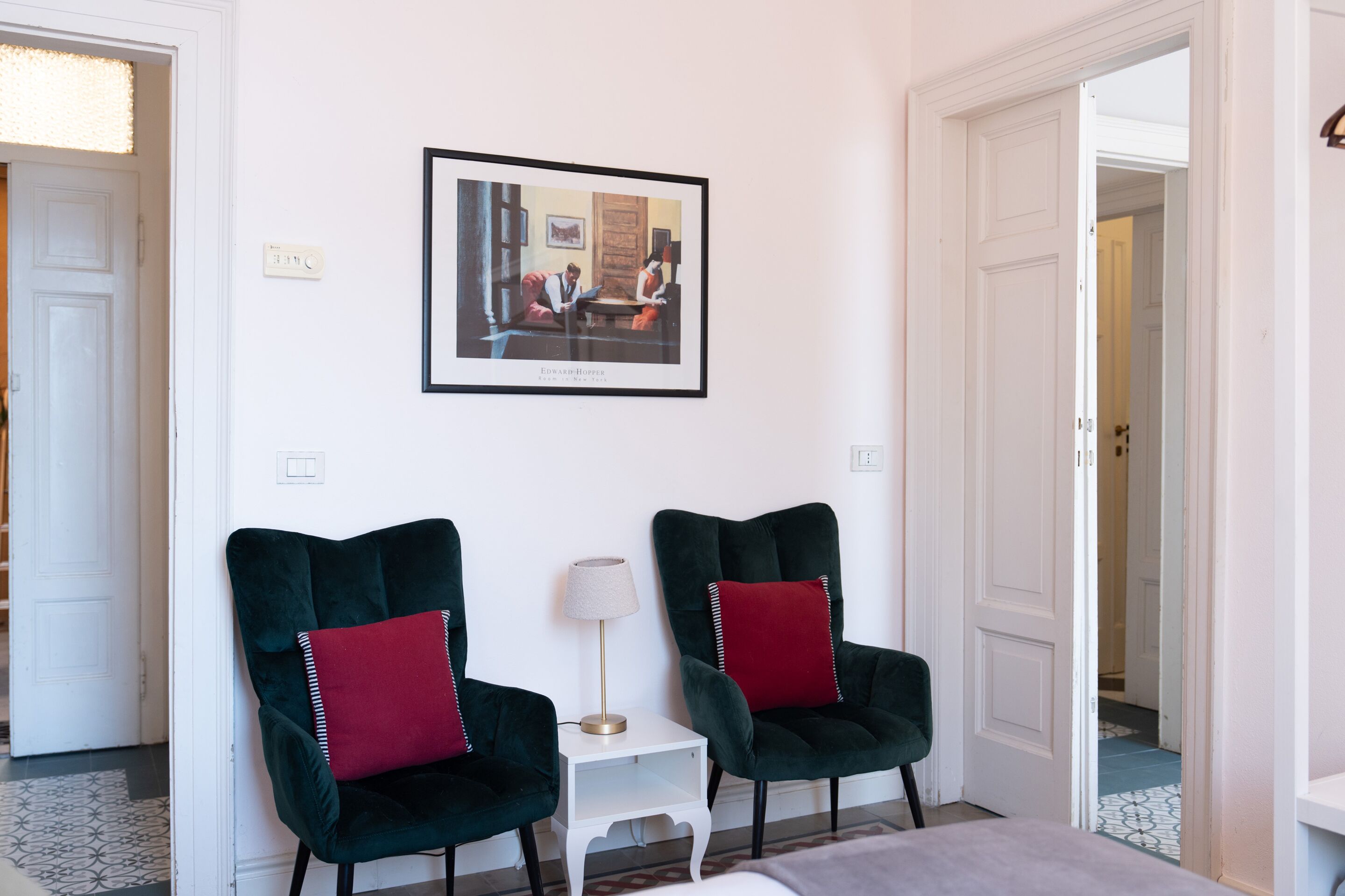 Een kamer met twee donkergroene fauteuils met rode kussens, een klein witte bijzettafeltje met een lamp en een ingelijste foto aan de muur.