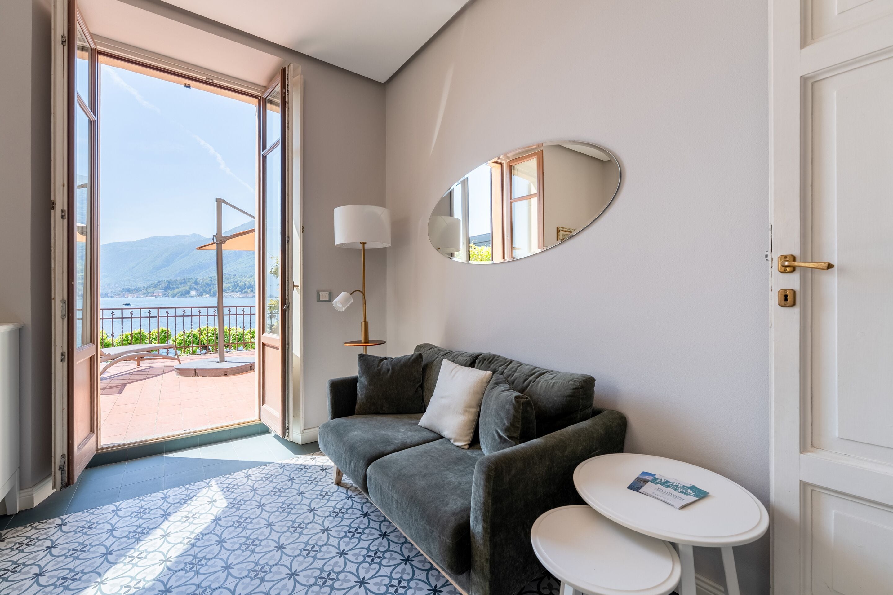 Una habitación con un sofá gris, una mesita blanca pequeña, un espejo redondo y vistas a un balcón con barandilla.