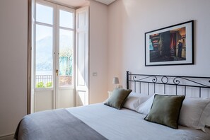 Une chambre à coucher avec un lit, des oreillers, une table de chevet, une lampe, un tableau encadré au mur et une fenêtre donnant sur un paysage verdoyant et des montagnes.