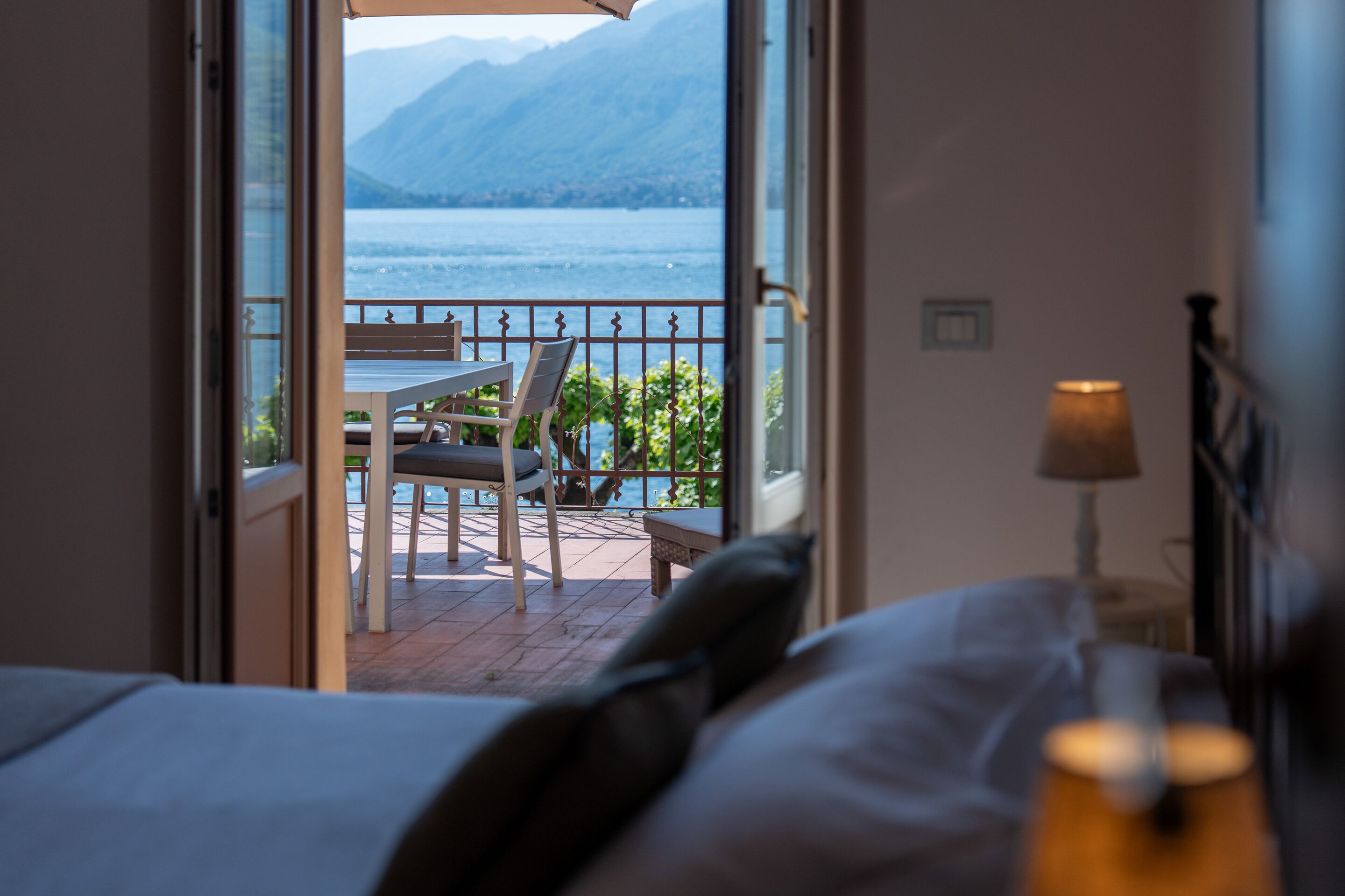 Habitación de hotel con una cama, una mesita de noche con lámpara y vistas a un balcón con mesa y sillas.
