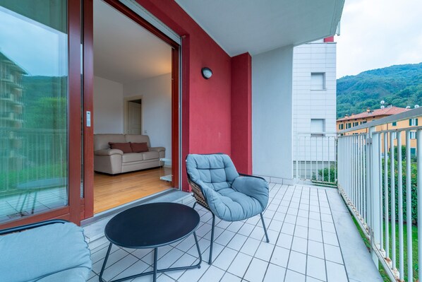 Un balcón con sofá y sillón, una puerta de vidrio que da a una habitación, y vistas a edificios y zonas verdes.