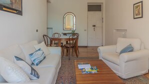 Ein Wohnzimmer mit einem weißen Sofa, einem Couchtisch mit Büchern, einem Esstisch mit Stühlen, einem Spiegel und einer Tür.